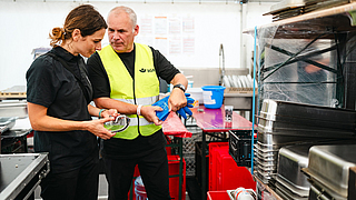 BGN-Arbeitsschutzexperte Stephan Thomassohn überprüft gemeinsam mit Clara Seipp von Sunny Side Catering die Persönliche Schutzausrüstung im Küchenzelt.