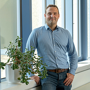 Dr. Andre Becker steht lächelnd vor einem großen Fenster, neben einer Pflanze auf der Fensterbank.