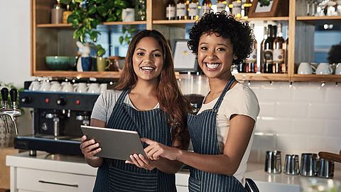 Zwei Frauen arbeiten im Bistro hinterm Tresen und schauen auf ein Tablet.