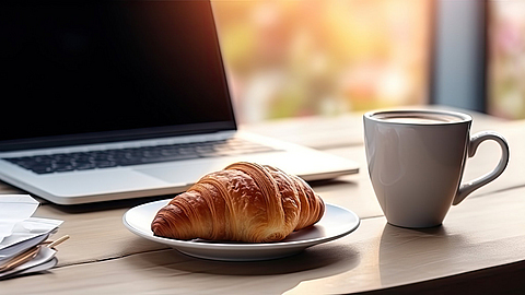 Frisches, leckeres Croissant und eine Tasse Kaffee vor dem Hintergrund des Arbeitsplatzes