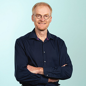 BGN-Mitarbeiter Dr. Peter Rietschel lächelt mit verschränkten Armen in die Kamera und steht vor hellblauem Hintergrund.