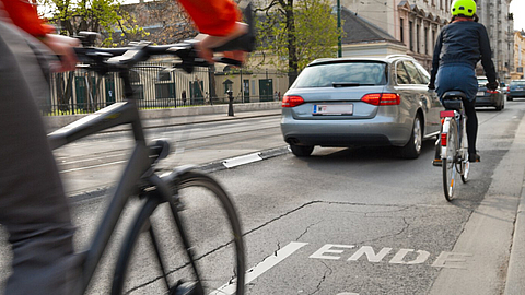Blick in eine Straße mit zwei Fahrradfahrern und einem Auto von hinten.