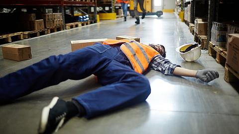 Gestürzter Arbeiter liegt auf dem Boden im Gang zwischen Lagerregalen