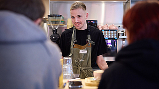Ein stets freundlicher Empfang: Mitarbeiter Simon Grünig begrüßt Gäste am Tresen. | © Dominik Buschardt Simon Grünig im Gespräch mit Gästen bei Coffee, Brownies and Downies
