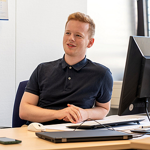 Jonas Brenneis sitzt an einem Schreibtisch vor einem Monitor und lächelt leicht, während er entspannt in die Ferne schaut
