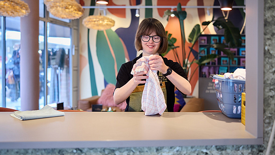 Immer mit einem Lächeln: „Ich freue mich auf meine Schicht am Samstag!“, sagt Mitarbeiterin Luise Brüggemann. | © Dominik Buschardt Luise Brüggemann faltet Wäsche im Inklusionscafé Coffee, Brownies and Downies