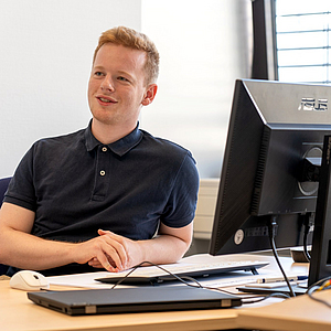 Jonas Brenneis sitzt an einem Schreibtisch vor einem Monitor und lächelt leicht, während er entspannt in die Ferne schaut