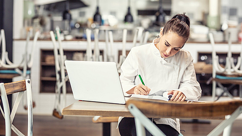 Die Restaurantköchin sitzt nach Feierabend am Tisch und erledigt ihre Buchhaltung vor einem offenen Laptop | © weyo - stock.adobe.com Die Restaurantköchin sitzt nach Feierabend am Tisch und erledigt ihre Buchhaltung vor einem offenen Laptop