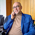 Herr Neuwerth trägt eine Brille und ein blaues Sakko, sitzt in einem modernen Büro und telefoniert mit einem Smartphone