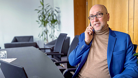 Herr Neuwerth trägt eine Brille und ein blaues Sakko, sitzt in einem modernen Büro und telefoniert mit einem Smartphone | © BGN/Saskia Nagel Herr Neuwerth trägt eine Brille und ein blaues Sakko, sitzt in einem modernen Büro und telefoniert mit einem Smartphone