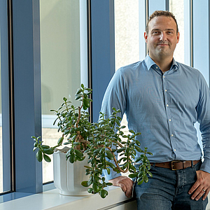 Dr. Andre Becker steht lächelnd vor einem großen Fenster, neben einer Pflanze auf der Fensterbank.