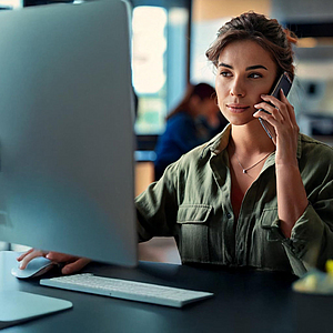 Eine junge Frau sitzt an ihrem Arbeitsplatz am Rechner und telefoniert