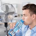 Mann trinkt Wasser im Büro mit Ventilator im Hintergrund