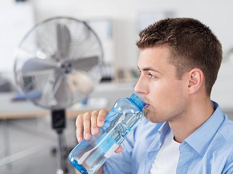 Mann trinkt Wasser im Büro mit Ventilator im Hintergrund