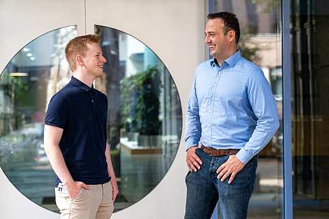 Von links: Jonas Brenneis und Dr. Andre Becker stehen sich lachend vor der Eingangstür des BGN-Schulungszentrums gegenüber